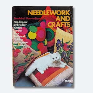 Puede incluir: Portada de libro vintage "Needlework and Crafts" con un gato blanco sobre un cojín colorido. La portada muestra proyectos de costura, bordado y acolchado. Es un libro de Simplicity's How-to Book.