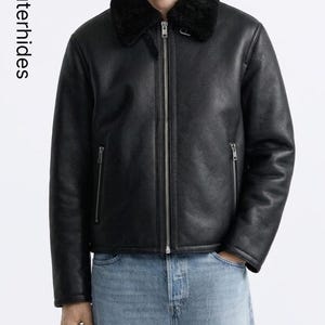 Puede incluir: Chaqueta de cuero negro con cuello de borrego negro y cremallera plateada. La chaqueta tiene dos bolsillos con cremallera en el pecho y dos en los laterales. El texto "Outerhides" está impreso verticalmente a la izquierda.