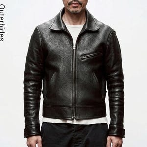 Puede incluir: Una chaqueta de cuero negro texturizado con cuello en punta, cremallera frontal completa y un bolsillo inclinado con cremallera en el pecho. La palabra Outerhides está impresa verticalmente en el lado izquierdo. La chaqueta tiene mangas largas.