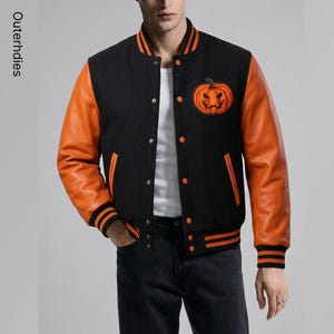 Op de afbeelding: Een zwart met oranje varsity-jack met een pompoenpatch op de borst. Het jack heeft lange oranje mouwen, een zwarte body, een drukknoopsluiting aan de voorzijde en gestreepte geribbelde boorden bij de mouwen en kraag. De tekst Outerhdies is zichtbaar.