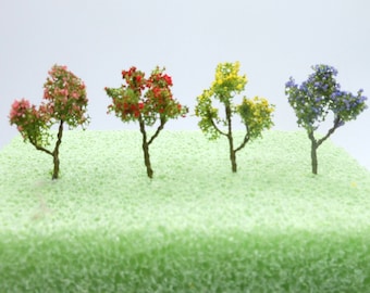 May include: Four miniature trees with different coloured flowers. The trees are on a green foam base. The trees are red, yellow, blue, and pink.