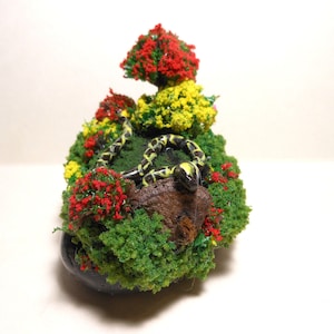 May include: A miniature garden scene with a black and yellow snake curled on a rock, surrounded by green moss, red and yellow flowers, and small trees.