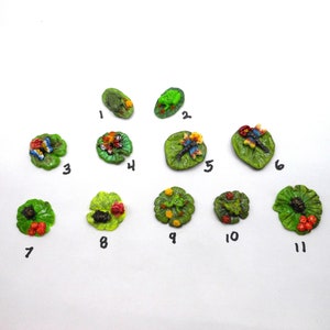 May include: Eleven miniature ceramic lily pads with various insects and frogs. The lily pads are green with brown edges and have a variety of colorful insects and frogs on them. The lily pads are numbered 1 through 11.