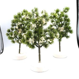 May include: Three miniature artificial trees with green foliage and white accents. The trees have brown trunks and are set on white circular bases. The background is white.