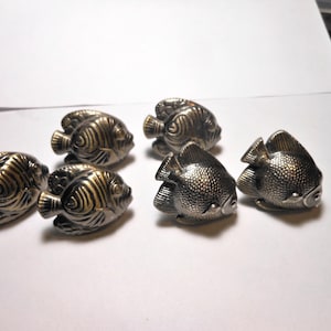 May include: Six decorative fish-shaped cabinet knobs with a metallic finish. The knobs are detailed with scales and fins, and are arranged on a white surface. The knobs are approximately 2 inches long.