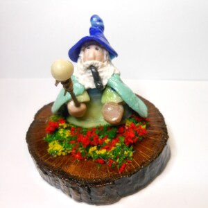 Può includere: Una statuetta in ceramica di un mago con un cappello blu, una barba bianca e una pipa. Tiene in mano un bastone con una sfera bianca in cima. La statuetta è seduta su una base di legno con fiori verdi e rossi.