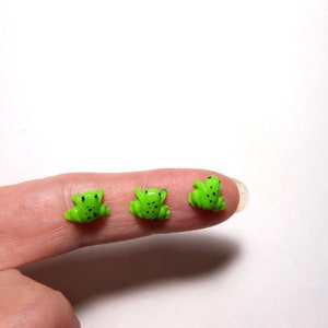May include: Three small green plastic frogs with black spots. The frogs are sitting on a finger.