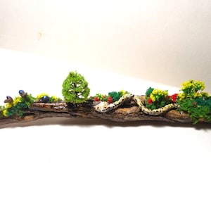 May include: A miniature forest scene with a small tree, flowers, and a snake on a piece of driftwood.
