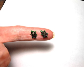 May include: A pair of small, black and gold turtle earrings with green accents. The earrings are shown on a finger.