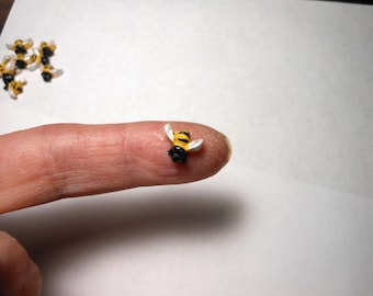 May include: Miniature bee figurine with yellow and black stripes, white wings, and a black body. Several other bee figurines are scattered in the background. The bee is resting on a finger.