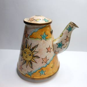 May include: A white ceramic teapot with a yellow, orange, and blue sun and star design. The teapot has a rounded body and a spout on the side.