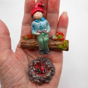 May include: A miniature figurine of a person wearing a red hat and blue jacket, sitting on a log with red flowers and a small hedgehog. A miniature campfire is below the log.
