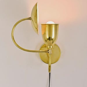 May include: A brass wall sconce with a curved arm and a half-dome shade. The sconce has a circular base and a cylindrical light fixture. A black cord extends from the bottom. The light is on.
