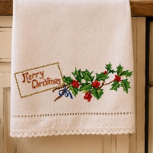 Puede incluir: Un paño de cocina blanco con un diseño de punto de cruz que muestra una etiqueta rectangular con el texto Merry Christmas en letras marrones, acompañado por un racimo de hojas de acebo verdes y bayas rojas atadas con un pequeño lazo azul.