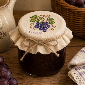 May include: A glass jar filled with dark purple jam, topped with a beige fabric cover featuring a cross-stitched design of purple grapes, green leaves, and the word Grape in purple text, secured with a tied brown twine bow around the rim.