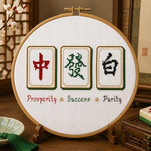 Puede incluir: Un bastidor de bordado circular con tela Aida blanca y un diseño de punto de cruz de tres fichas de Mahjong. Las fichas muestran caracteres en rojo, verde y negro. Debajo de las fichas, las palabras Prosperity, Success y Purity están bordadas.