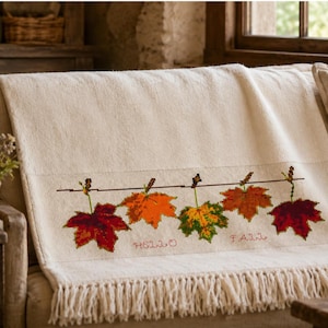 Può includere: Una coperta color crema con un disegno a punto croce di cinque foglie autunnali colorate appese a una linea orizzontale. Il testo "HELLO FALL" è ricamato in rosso sotto le foglie e il bordo inferiore presenta lunghe frange.