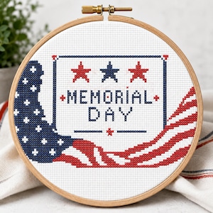 May include: A cross-stitch embroidery hoop featuring a patriotic design for Memorial Day. The design includes the text "MEMORIAL DAY" in blue, red stars, and a waving American flag. The embroidery is set in a wooden hoop.