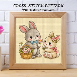 May include: A framed cross-stitch pattern featuring two adorable bunnies. One gray bunny wears a blue bow tie and holds flowers, while the other, a beige bunny, has a flower crown and pink scarf. A basket of flowers sits beside the gray bunny. The text "CROSS-STITCH PATTERN PDF Instant Download" is at the top.