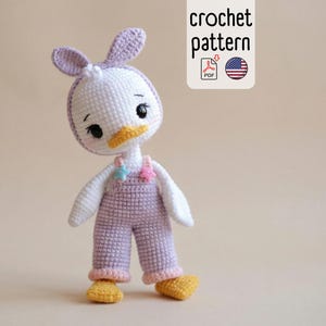Puede incluir: Una muñeca de pato de ganchillo hecha a mano que lleva un mono lavanda y pies amarillos. El pato tiene una cabeza blanca con orejas lavanda y un pico amarillo. La imagen incluye un rectángulo blanco con las palabras "crochet pattern".