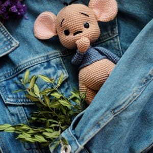 May include: A crocheted doll with a light brown body, pink ears, and a blue top, nestled in a blue denim jacket pocket. The doll has black eyes and a contemplative look. Green leaves and dried flowers provide a natural contrast.
