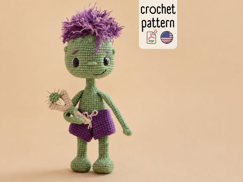 May include: A green crocheted doll with messy purple hair, wearing purple shorts and holding a small beige slingshot. The doll has black button eyes and a stitched smile. A white rectangular label displays the text crochet pattern.