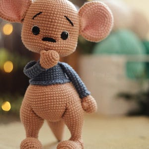 May include: A handmade crochet kangaroo doll in shades of tan and pink, wearing a blue jumper. The doll has large, round ears, black eyes, and a thoughtful expression. The toy is standing on a textured surface.