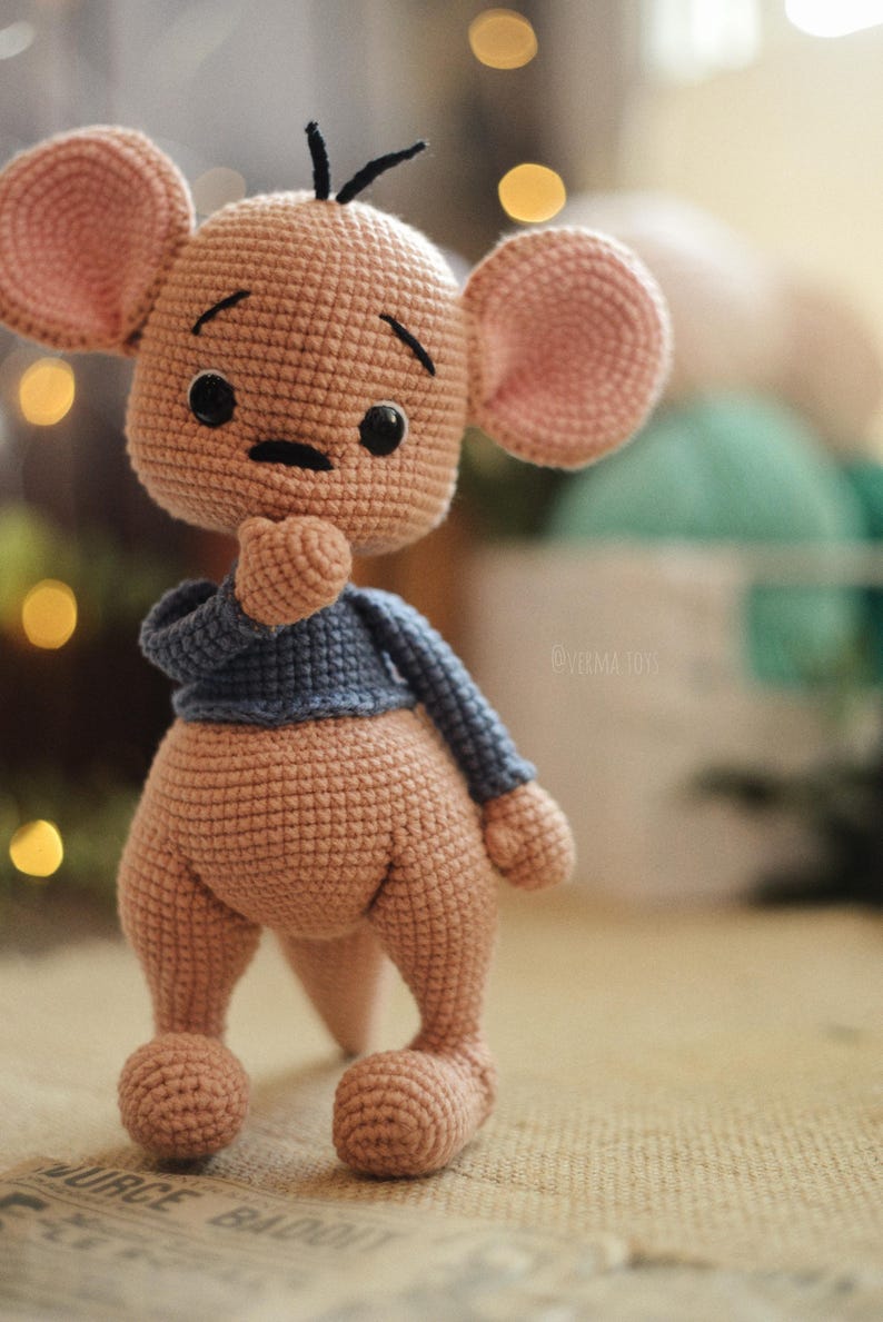 May include: A handmade crochet kangaroo doll in shades of tan and pink, wearing a blue jumper. The doll has large, round ears, black eyes, and a thoughtful expression. The toy is standing on a textured surface.