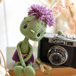 May include: A green crocheted doll with a large head, black button eyes, and messy purple yarn hair. The doll wears a purple skirt and sits next to a black vintage camera with the text "Смена-8" printed on the front of the camera body.