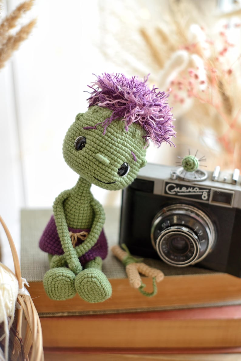 May include: A green crocheted doll with a large head, black button eyes, and messy purple yarn hair. The doll wears a purple skirt and sits next to a black vintage camera with the text "Смена-8" printed on the front of the camera body.