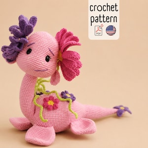 May include: A pink crocheted axolotl plush toy featuring purple and pink frilled gills, small embroidered eyes, and a long tail. The body is decorated with small purple and pink flowers and green vines. Text reads crochet pattern.
