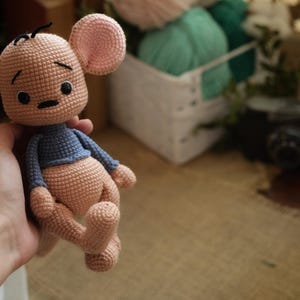 May include: A handmade crochet mouse doll with a light brown body, pink ears, and a blue top. The doll has black eyes and a small black nose. The doll is being held in a hand.