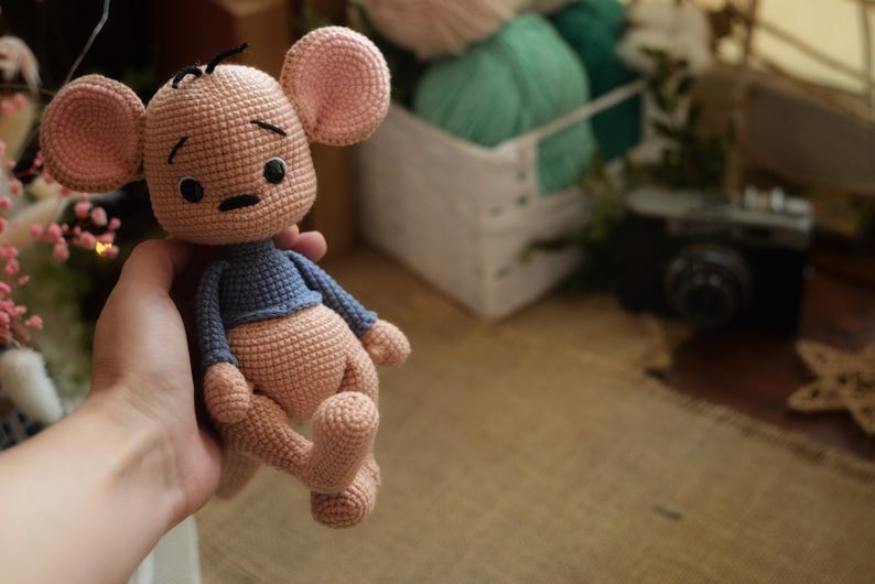 May include: A handmade crochet mouse doll with a light brown body, pink ears, and a blue top. The doll has black eyes and a small black nose. The doll is being held in a hand.