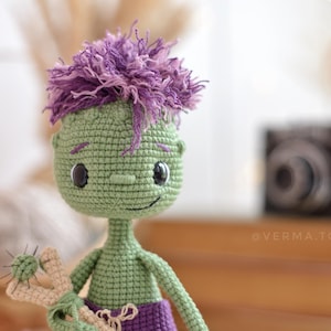 May include: A green crocheted doll with a large head, dark eyes, and a small nose. The doll has messy purple yarn hair, a slender neck, and wears purple trousers. It holds a beige slingshot with a small green ball. The text @VERMA.TOYS is visible.