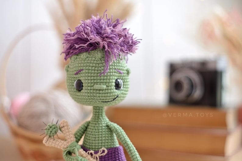 May include: A green crocheted doll with a large head, dark eyes, and a small nose. The doll has messy purple yarn hair, a slender neck, and wears purple trousers. It holds a beige slingshot with a small green ball. The text @VERMA.TOYS is visible.