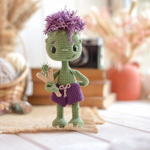 May include: A green crocheted doll with a large head, purple hair, and purple shorts. The doll has black button eyes, a small stitched smile, and is holding a small beige and green crocheted slingshot accessory in its right hand.