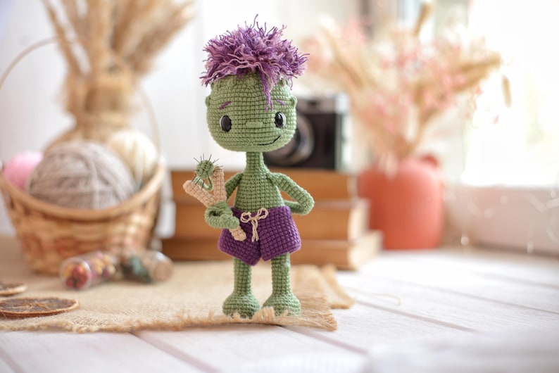 May include: A green crocheted doll with a large head, purple hair, and purple shorts. The doll has black button eyes, a small stitched smile, and is holding a small beige and green crocheted slingshot accessory in its right hand.