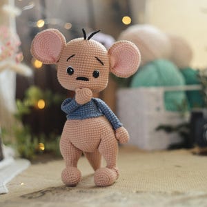 May include: A handmade crochet mouse doll in a standing pose. The doll has a light brown body, large pink ears, and a blue jumper. The mouse is holding its chin with one hand, and has black eyes and a small black nose.