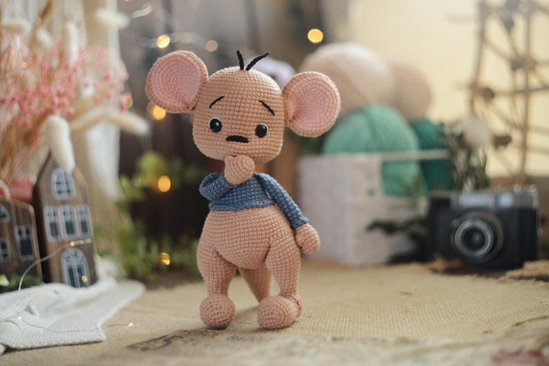 May include: A handmade crochet mouse doll in a standing pose. The doll has a light brown body, large pink ears, and a blue jumper. The mouse is holding its chin with one hand, and has black eyes and a small black nose.