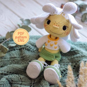 May include: A crocheted doll with a yellow head, freckles, and a white flower-shaped hat. It wears a white long-sleeved shirt with yellow heart patterns, a green skirt, and white and green sneakers. A circular tag reads PDF pattern ENG.