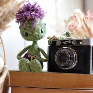 May include: A green crocheted doll with purple hair and purple shorts sitting next to a black vintage camera with the text Smena-8 printed on the front. The doll has large black eyes and a small stitched smile, crafted with detailed yarn texture.