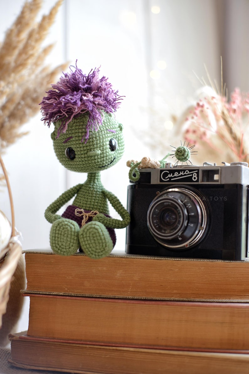 May include: A green crocheted doll with purple hair and purple shorts sitting next to a black vintage camera with the text Smena-8 printed on the front. The doll has large black eyes and a small stitched smile, crafted with detailed yarn texture.