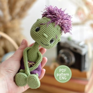 May include: A small, handmade crochet doll with green skin, large black eyes, and a tuft of purple hair. The doll wears purple shorts and has long, thin arms and legs. A circular green graphic on the image contains the text PDF pattern ENG.
