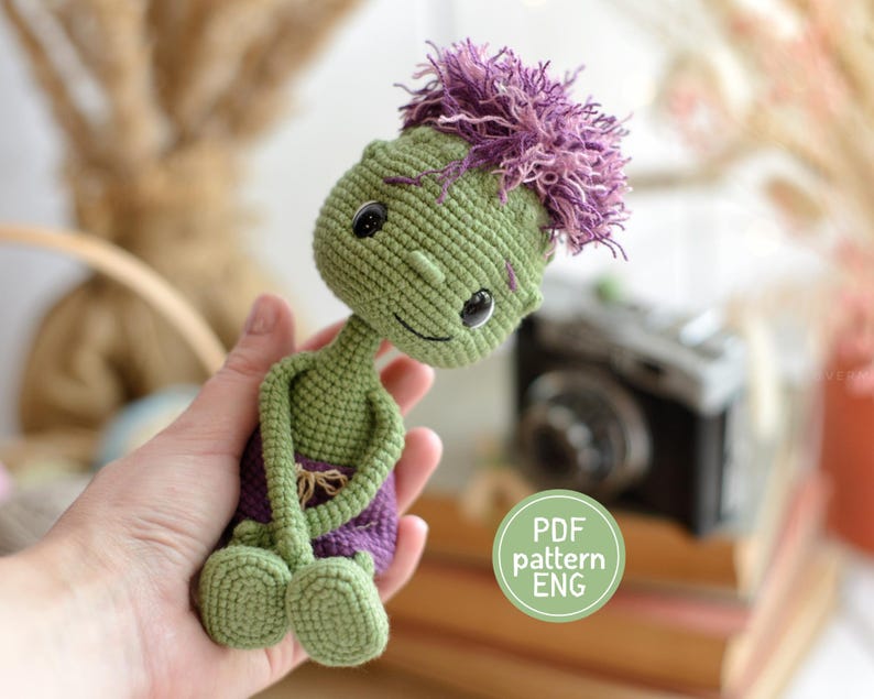 May include: A small, handmade crochet doll with green skin, large black eyes, and a tuft of purple hair. The doll wears purple shorts and has long, thin arms and legs. A circular green graphic on the image contains the text PDF pattern ENG.