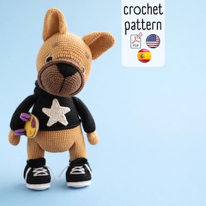 Op de afbeelding: Een gehaakt bulldog-speeltje in bruin, zwart en wit. De bulldog draagt een zwarte hoodie met een witte ster en zwart-witte sneakers. Hij houdt een klein skateboard vast. De afbeelding bevat de tekst "crochet pattern".