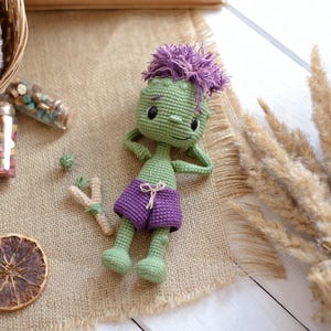 May include: A green crocheted doll with purple hair and purple shorts, lying down with hands behind its head. The doll features black embroidered eyes and a small smile, crafted with a textured yarn finish and a simple drawstring on the shorts.