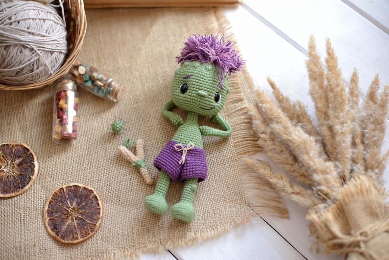 May include: A green crocheted doll with purple hair and purple shorts, lying down with hands behind its head. The doll features black embroidered eyes and a small smile, crafted with a textured yarn finish and a simple drawstring on the shorts.
