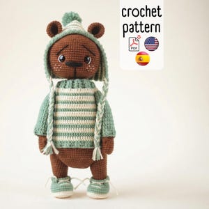 Puede incluir: Un oso de ganchillo marrón hecho a mano que lleva un suéter a rayas verdes y blancas, un gorro a juego y zapatillas deportivas. El oso tiene una expresión dulce y es un artículo hecho a mano encantador. La imagen incluye el texto "crochet pattern".