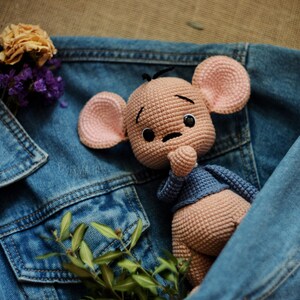 May include: A crocheted mouse doll with pink ears and a blue sweater is nestled in a denim jacket. Dried flowers and greenery add colour. The doll has a sad expression. The jacket is a classic blue.