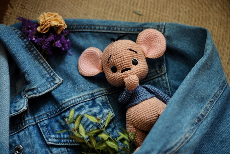 May include: A crocheted mouse doll with pink ears and a blue sweater is nestled in a denim jacket. Dried flowers and greenery add colour. The doll has a sad expression. The jacket is a classic blue.